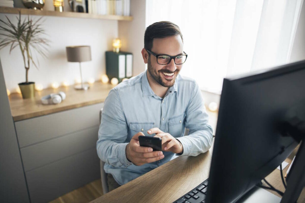 Smiling handsome freelancer working remotely from home. He is using phone.